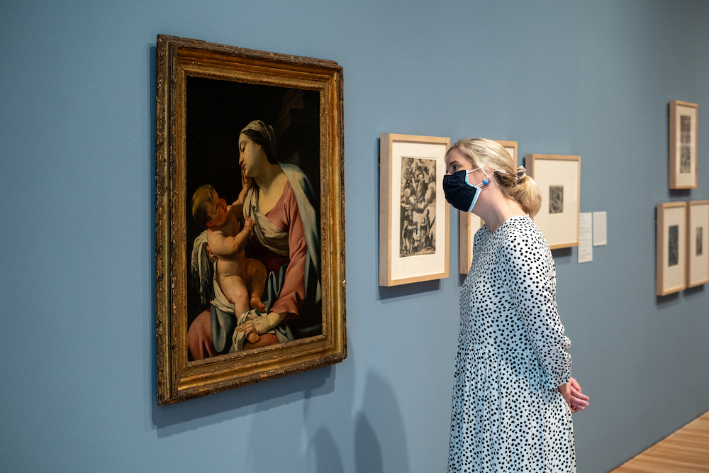 A masked woman inside a museum looking up at a work of art on the wall.