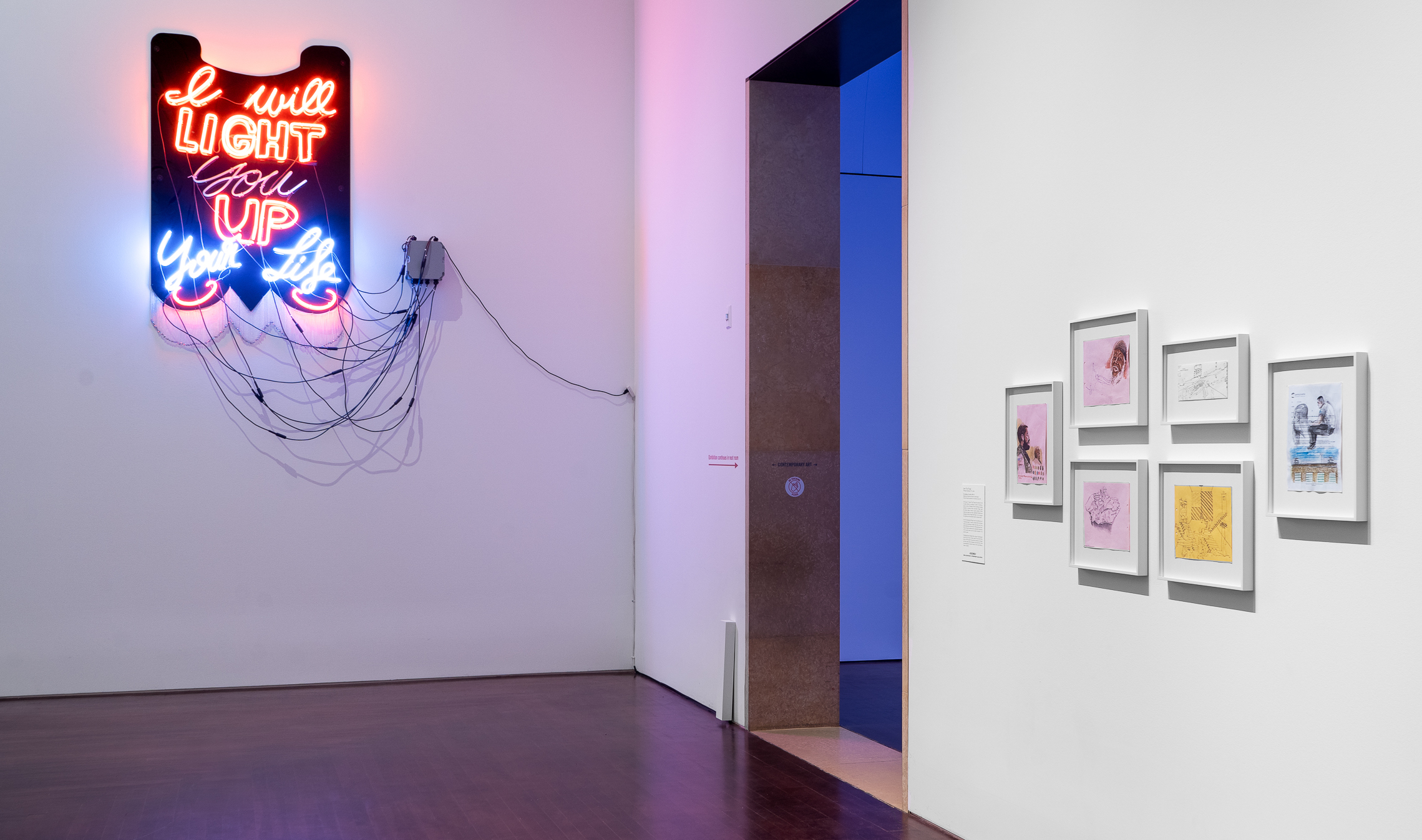 A view inside a gallery. A neon-lit work is positioned high on one wall with wires coming down. It alternates saying 