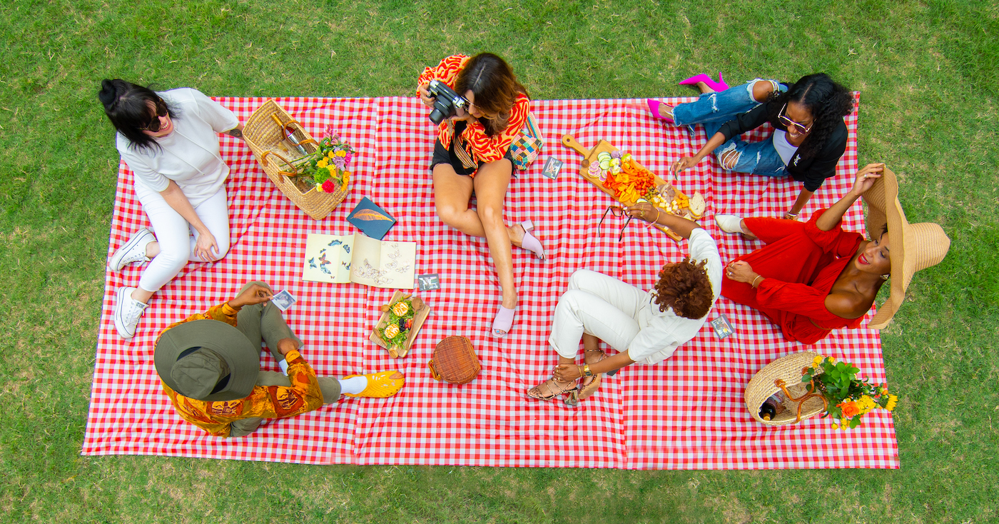 A group of people sat on a large picnic blanket. On the blanket are boards of food and baskets containing flowers and wine