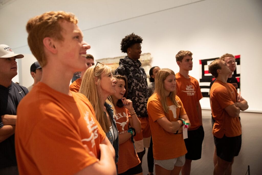 A group of burnt orange clad University of Texas at Austin students looking at artwork with a gallery instructor. 