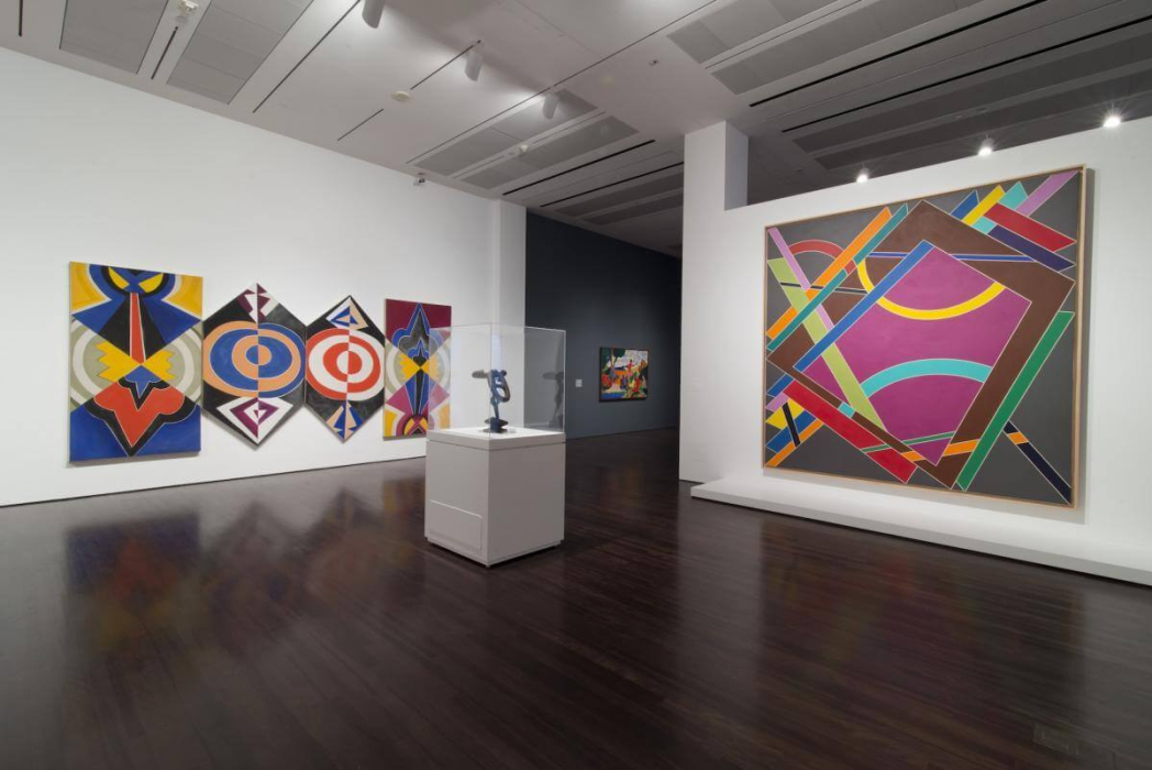 An art gallery featuring several abstract paintings and a sculpture. The left wall displays five colorful abstract paintings with geometric shapes and patterns. In the center of the room, a sculpture is showcased in a glass case on a white pedestal. The right wall has a large abstract painting with overlapping geometric shapes in various colors. The gallery has dark wooden floors and recessed lighting, creating a well-lit environment that highlights the artworks.