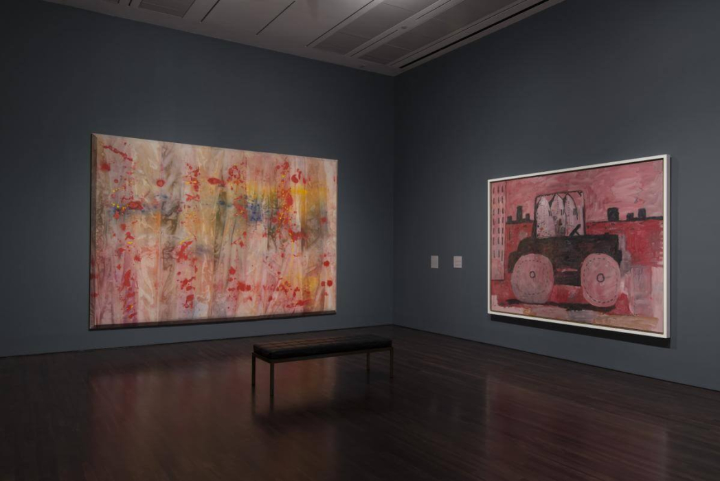 An art gallery with two large paintings displayed on the walls. The left painting is an abstract piece with vibrant colors, including red, yellow, blue, and green, creating a dynamic and chaotic effect. The right painting depicts a vehicle, possibly a tractor, in shades of red and black, set against a background with buildings. A bench in the center of the room offers a place for viewers to sit and observe the artwork. The gallery has dark walls and a wooden floor, highlighting the vibrant colors of the paintings.