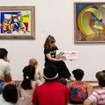 Storytime program. Person sitting in front of framed works of art on the wall of a gallery holding up a children's book. Children and adults sit on the ground facing the person reading.