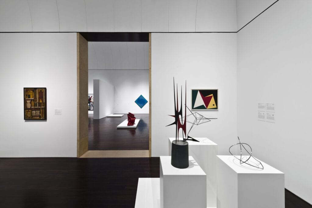 Interior art gallery space. In the foreground are several sculptural artworks on pedestals. In the background are paintings on a wall and more visible in the adjoining gallery space.