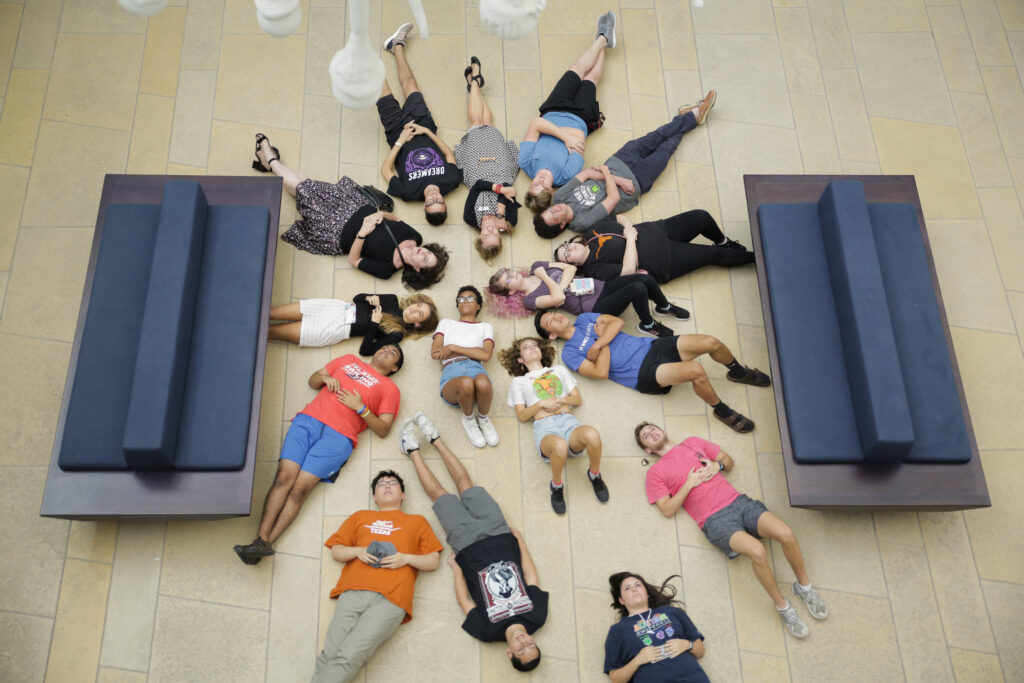 A group of young students and their gallery instructor lying on the museum floor looking up at an artwork.