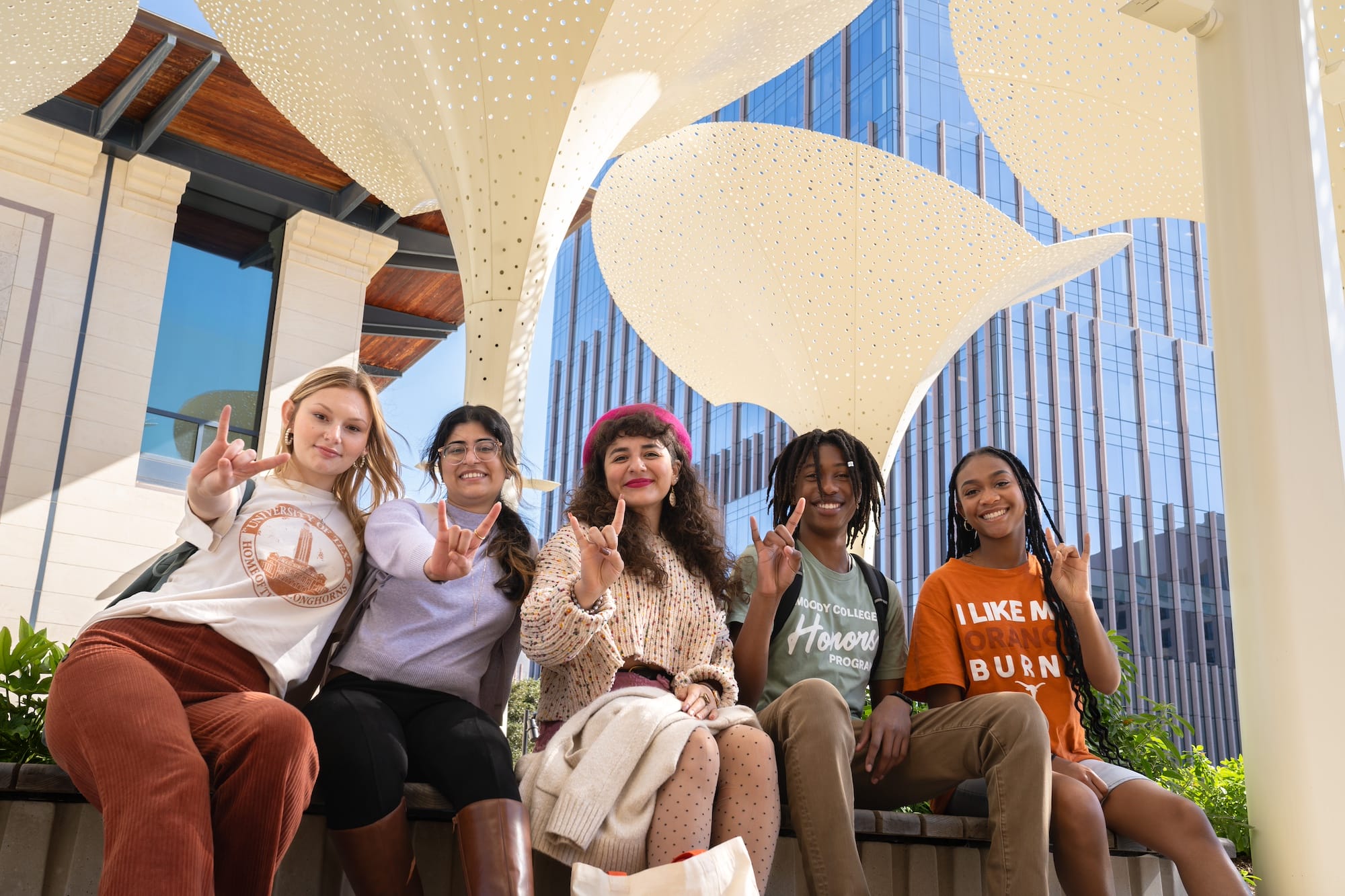 A group of students from the University of Texas at Austin holding up their hands using the "hook 'em" hand gesture. They are sitting on the grounds of the Blanton Museum of Art under tall petal-like structures