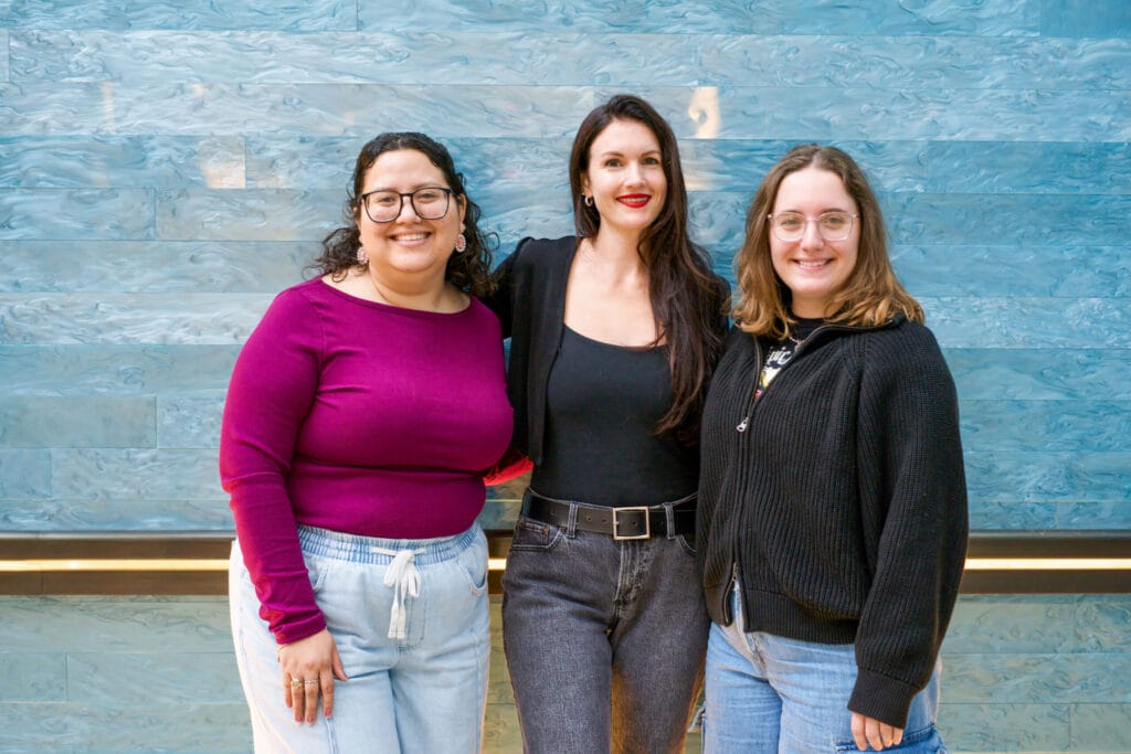 Three Blanton graduate fellows pose in front of Stacked Waters by Teresita Fernandez in the Blanton Museum atrium, a large artwork featuring blue glass