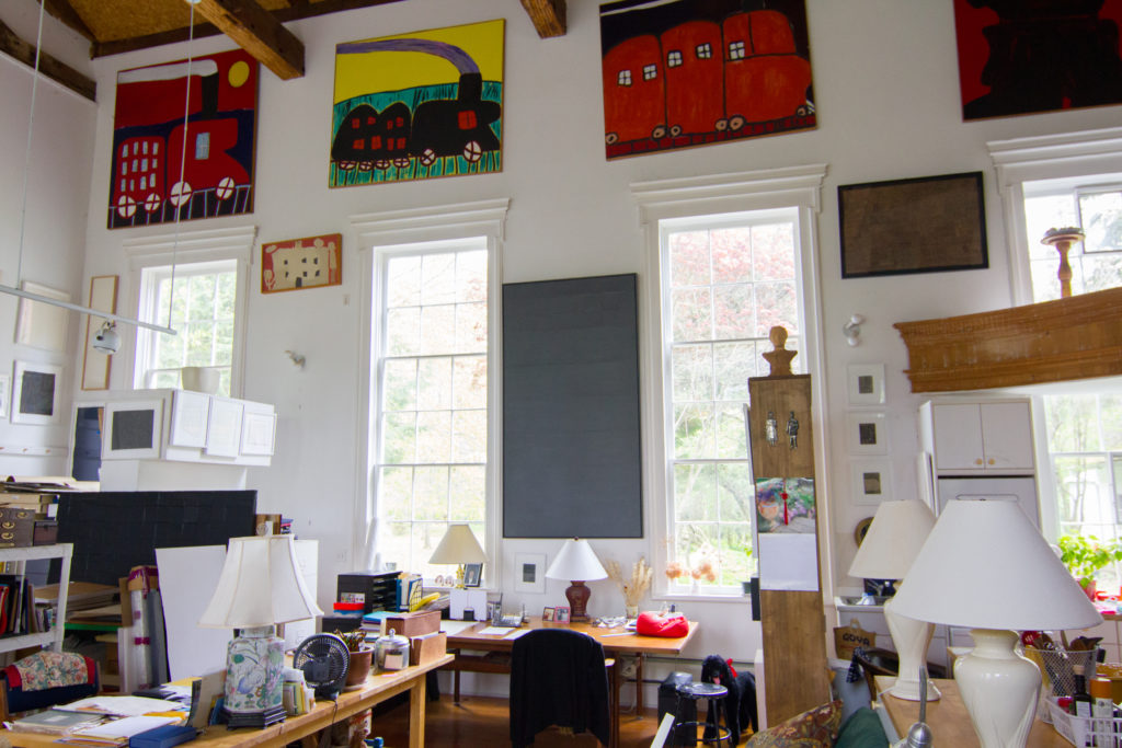 A room with a giant white wall with vertical windows spanning across it and various artworks hung on it. The rest of the room is filled with artworks/tools