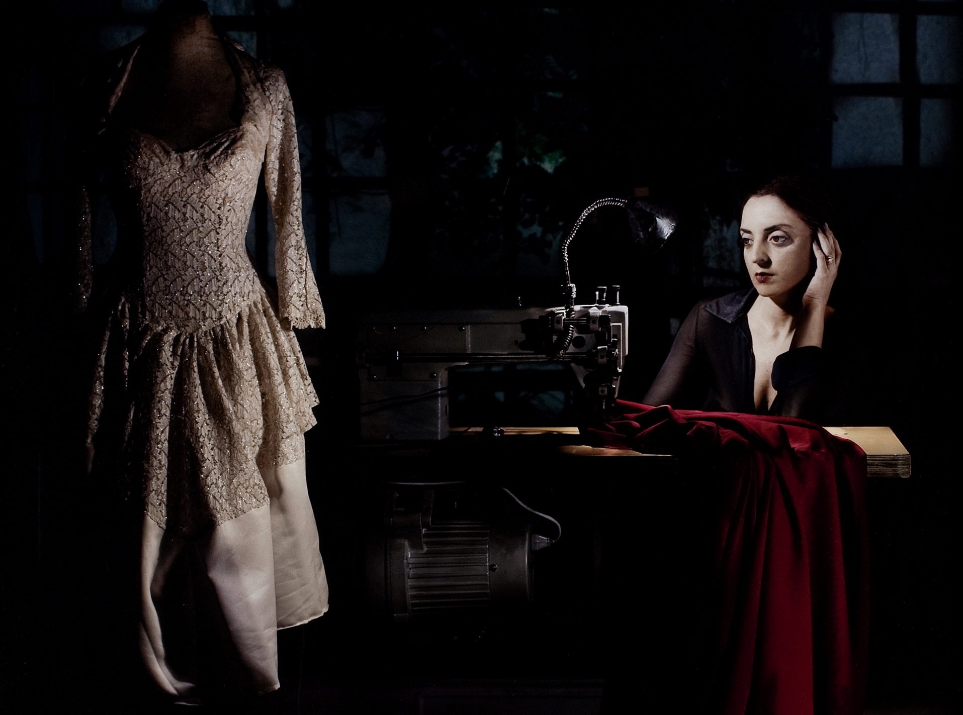 Nicola Costantino at sewing machine looking at a dress on a mannequin. Dark lighting creating a high contrast from Costantino's skin.