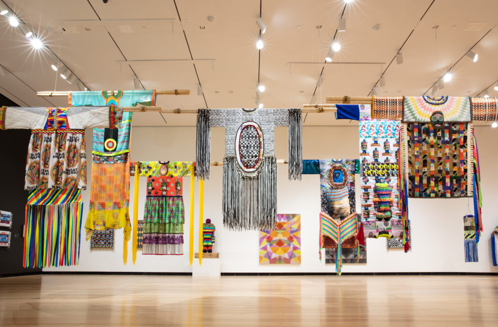 Installation view of colorful garments hanging from the ceiling shown in the exhibition Jeffrey Gibson: This is the Day