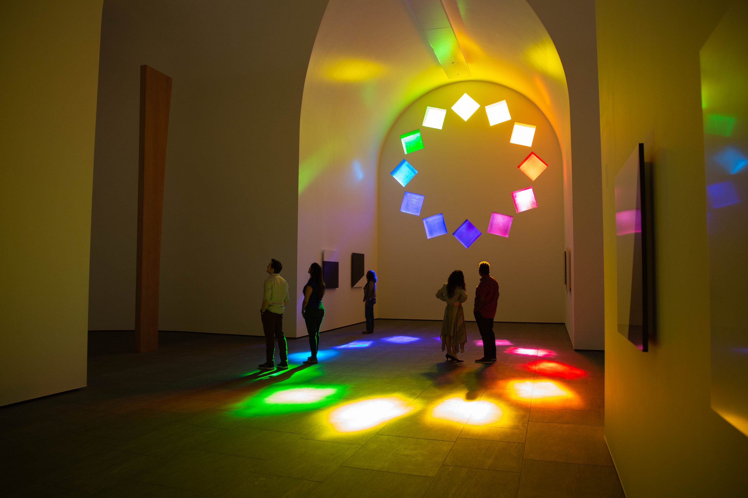 People standing inside a chapel-like building. A color wheel window of multi-colored glass squares tumbling in a circle project vivid colors on the floor of the building