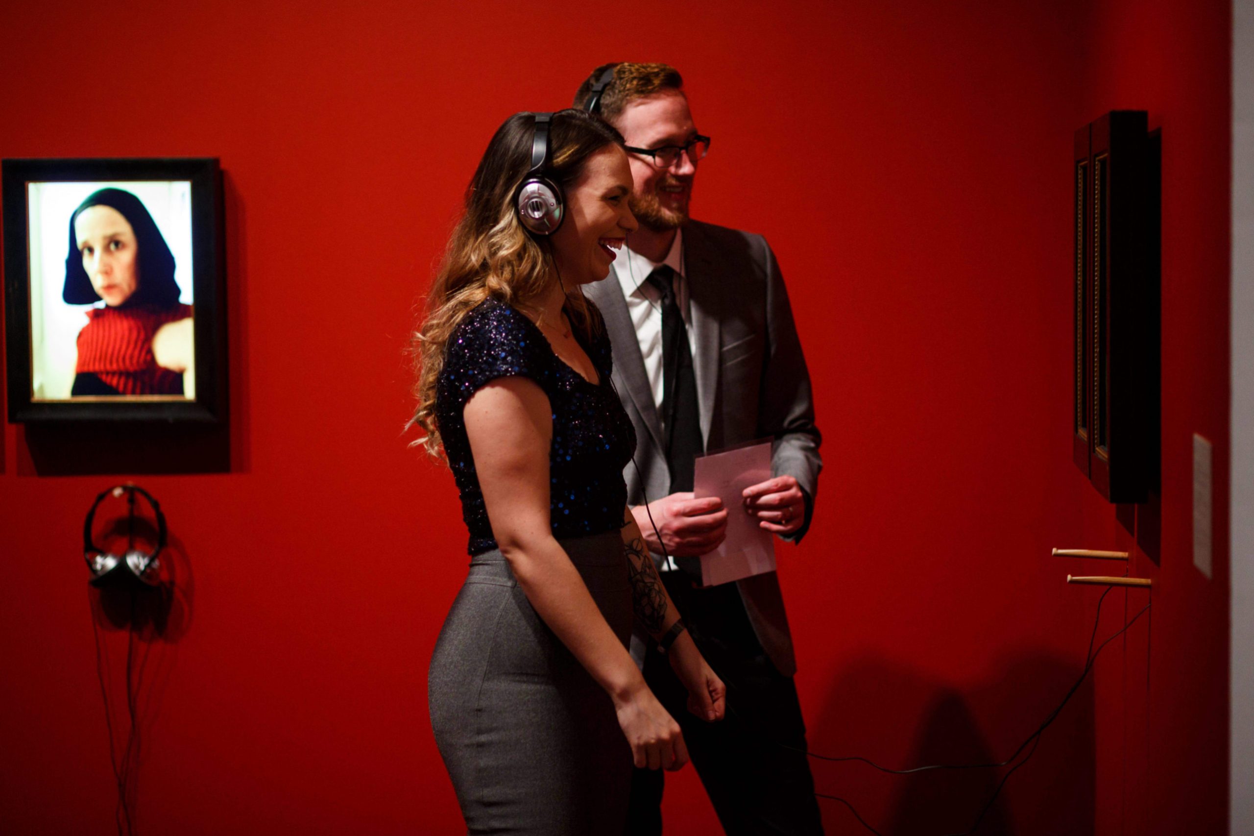 a cocktail attired couple listen to a nina katchadourian video through headphones and smile. The room is dark red and a still image of nina is present in the background.
