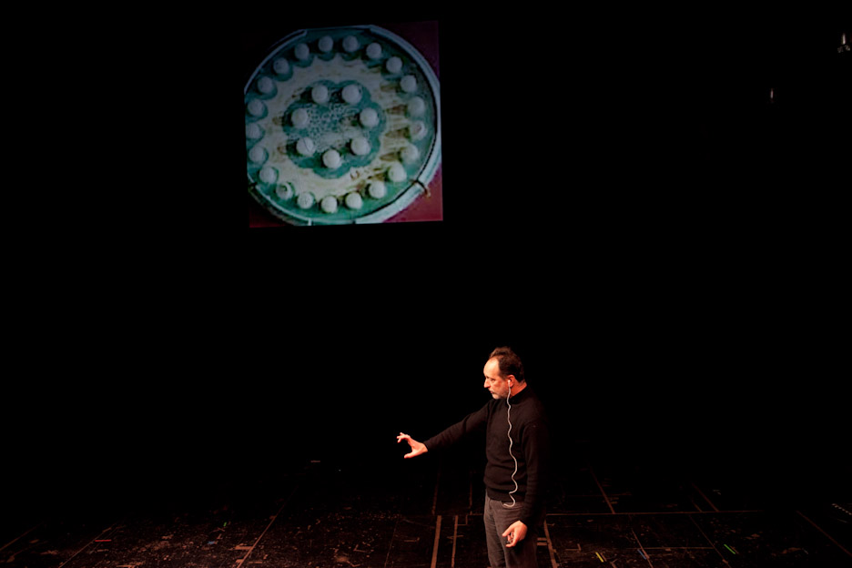 A man stands on a stage with an outstretched hand. He is wearing a black outfit and in the background there is a circular prop with white centric dots.
