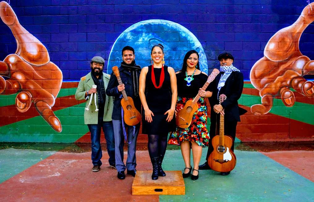 Musical group, Güateque Son, pose for a photo. Members hold their instruments. String instruments and guitars. They pose in front of a mural. The mural has two outstretched hands in between is a blue planet either the moon or earth. There is a green and red line pattern taking up half the mural. The pattern extends past the mural to the sidewalk where the musical group poses.