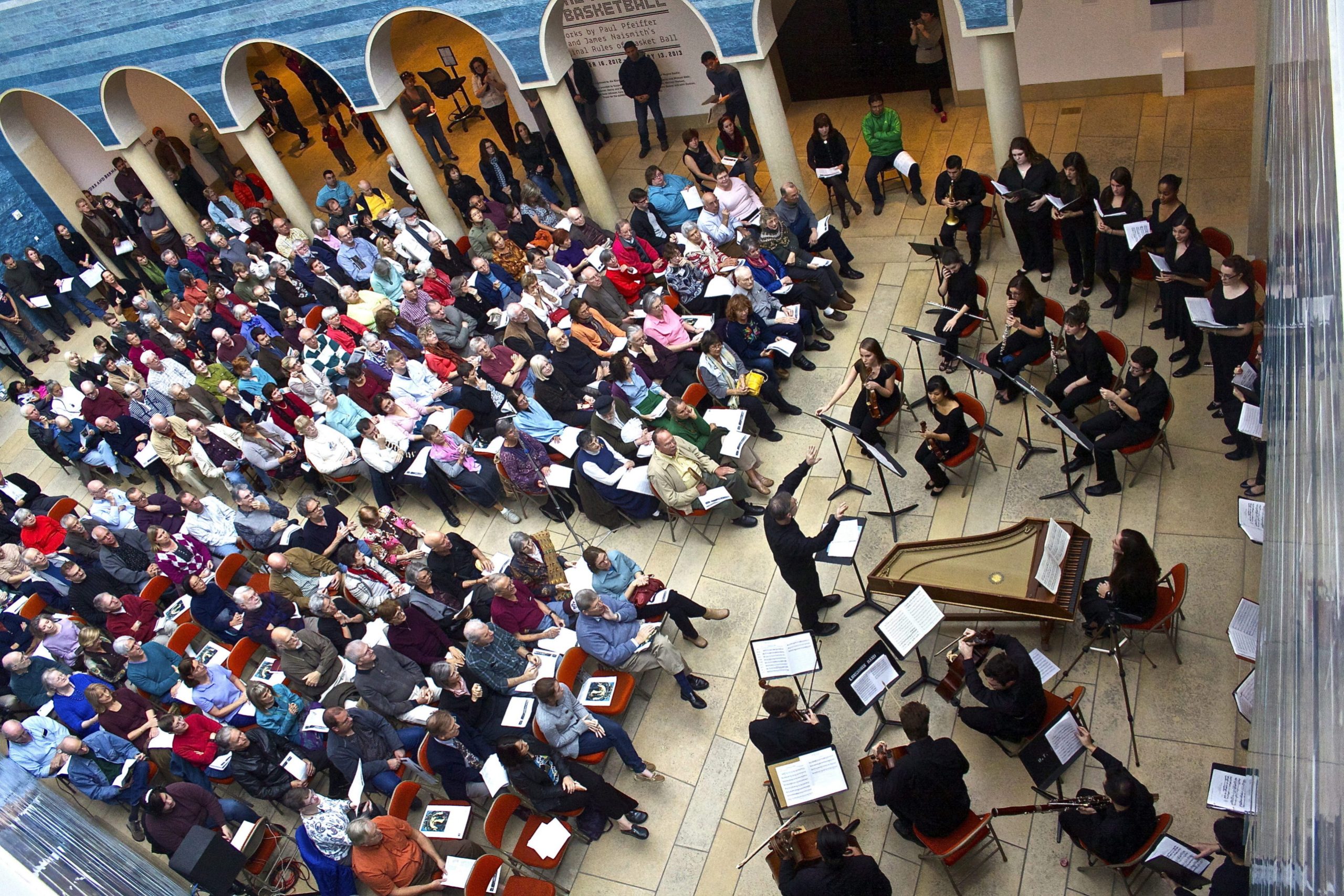 Photo of the atrium at the Blanton Museum of Art featuring a large audience sitting, listening to an orchestra perform. The guests are seated in rows on the left facing the orchestra on the right. The orchestra members are all wearing a coordinated black uniform playing a variety of instruments.