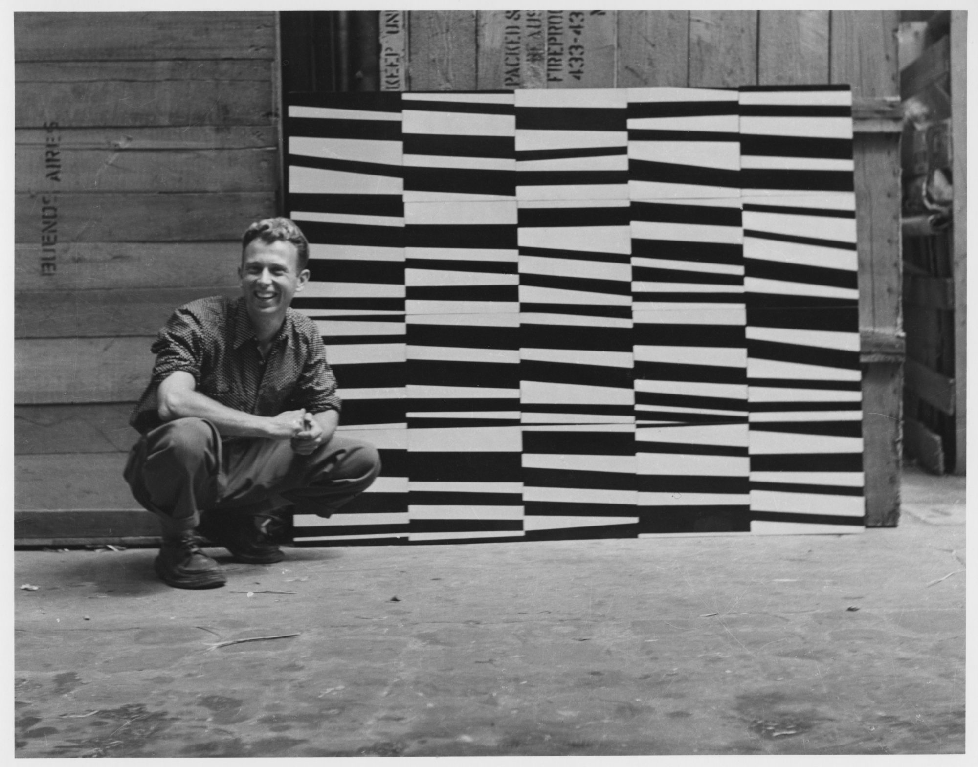 A photograph of the artist Ellsworth Kelly, crouching next to his upside down painting of patterns titled 