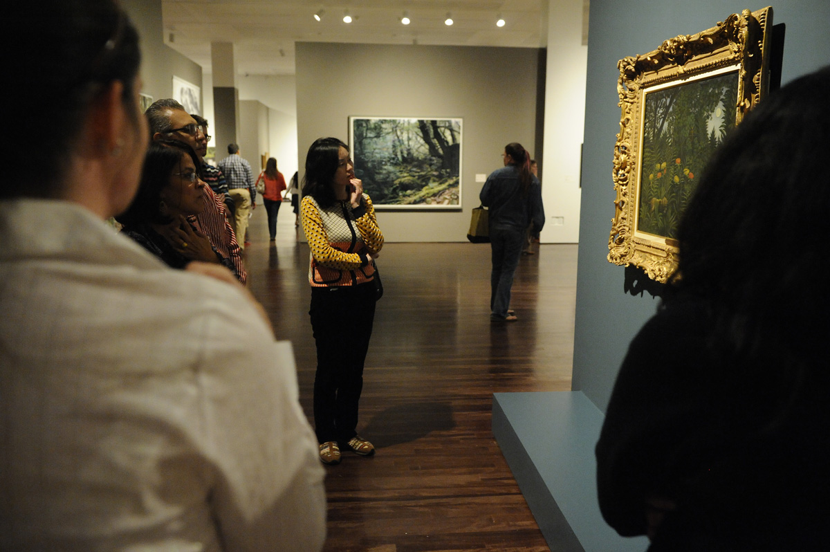 Six adults stand in front of a rousseau painting of a tropical forest in a large guilded frame. A larger than life contemporary landscape photograph can be seen in the distance.