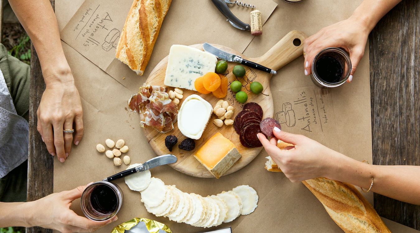 Various hands outstretched over a cheese board with crackers and bread scattered nearby.