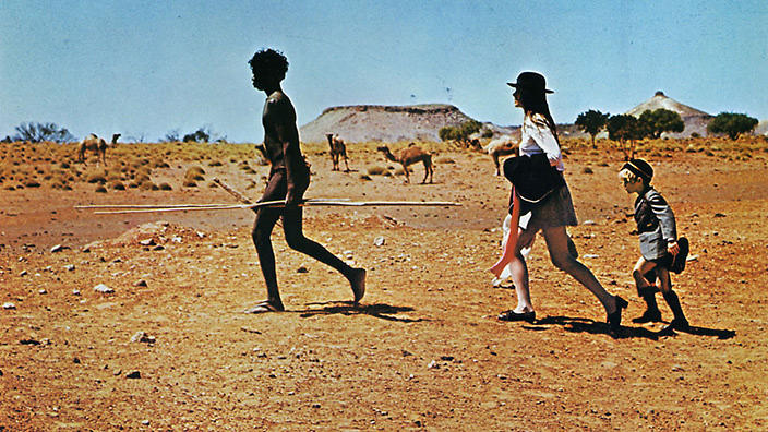 an barefoot indigenous australian leads a woman carrying a blazer dressed in pumps, skirt, blouse and hat and a child dressed in a school uniform-type outfit across a barren landscape with camels in the background