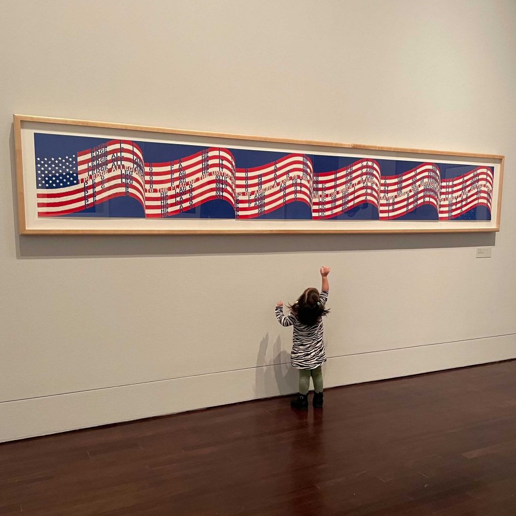 A toddler girl looking up and pointing at a long horizontal print of the USA flag hanging on the wall.