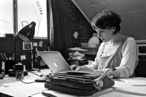 photo of Lenka Clayton at her desk working at her computer. On her desk is a typewriter, a lamp, and some other office items