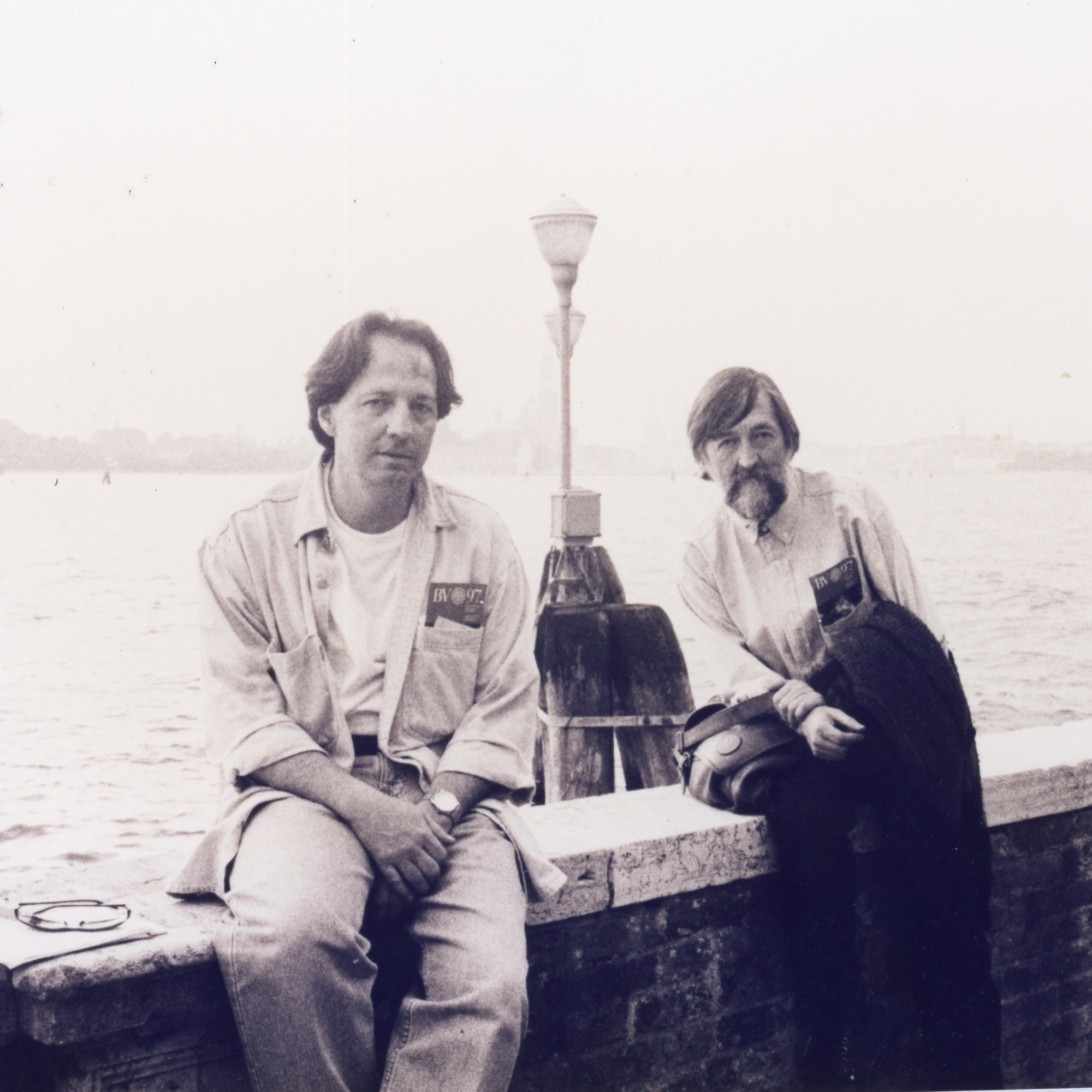 A black and white photo of two men sat on a wall that overlooks a body of water.