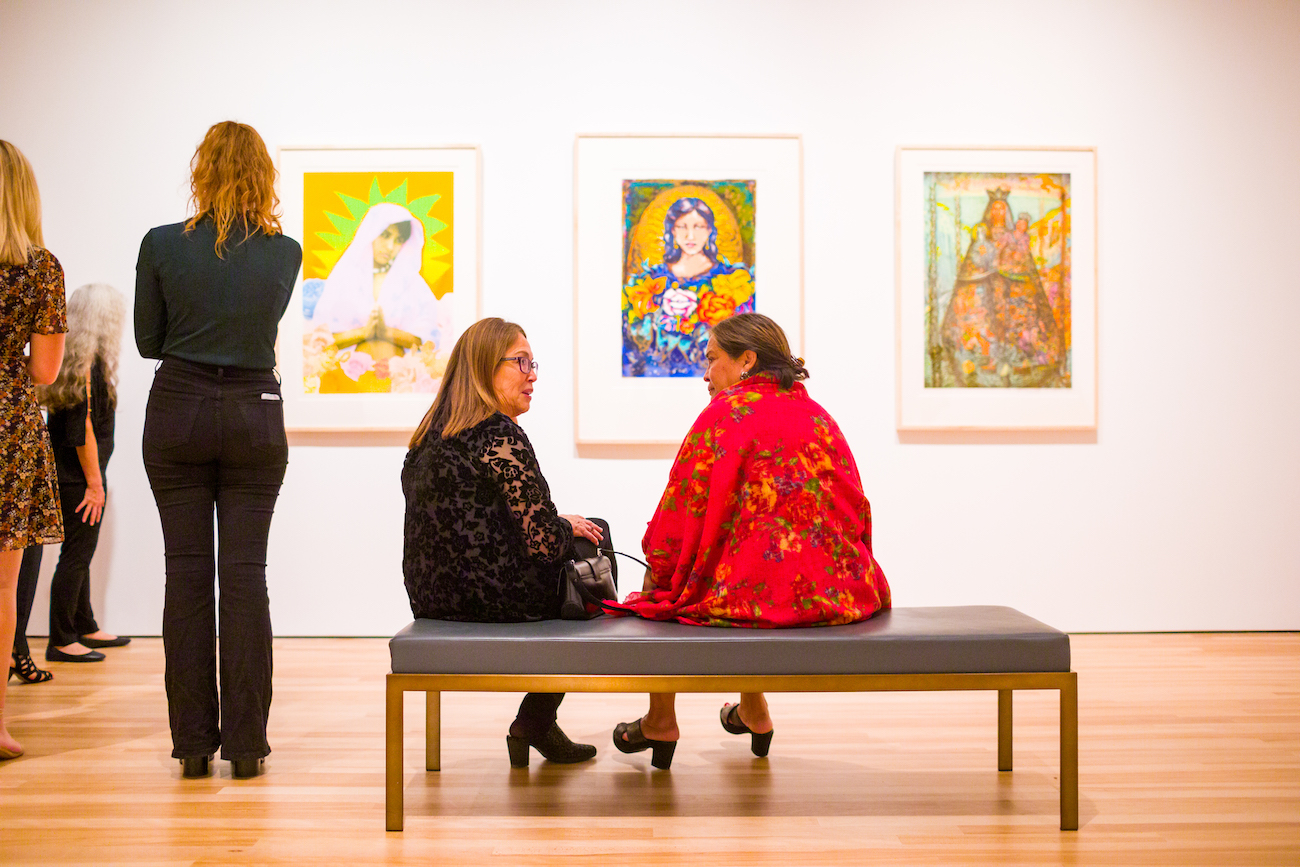 Two women sat on a bench in front of 3 framed prints with depictions of the Virgin and Child.