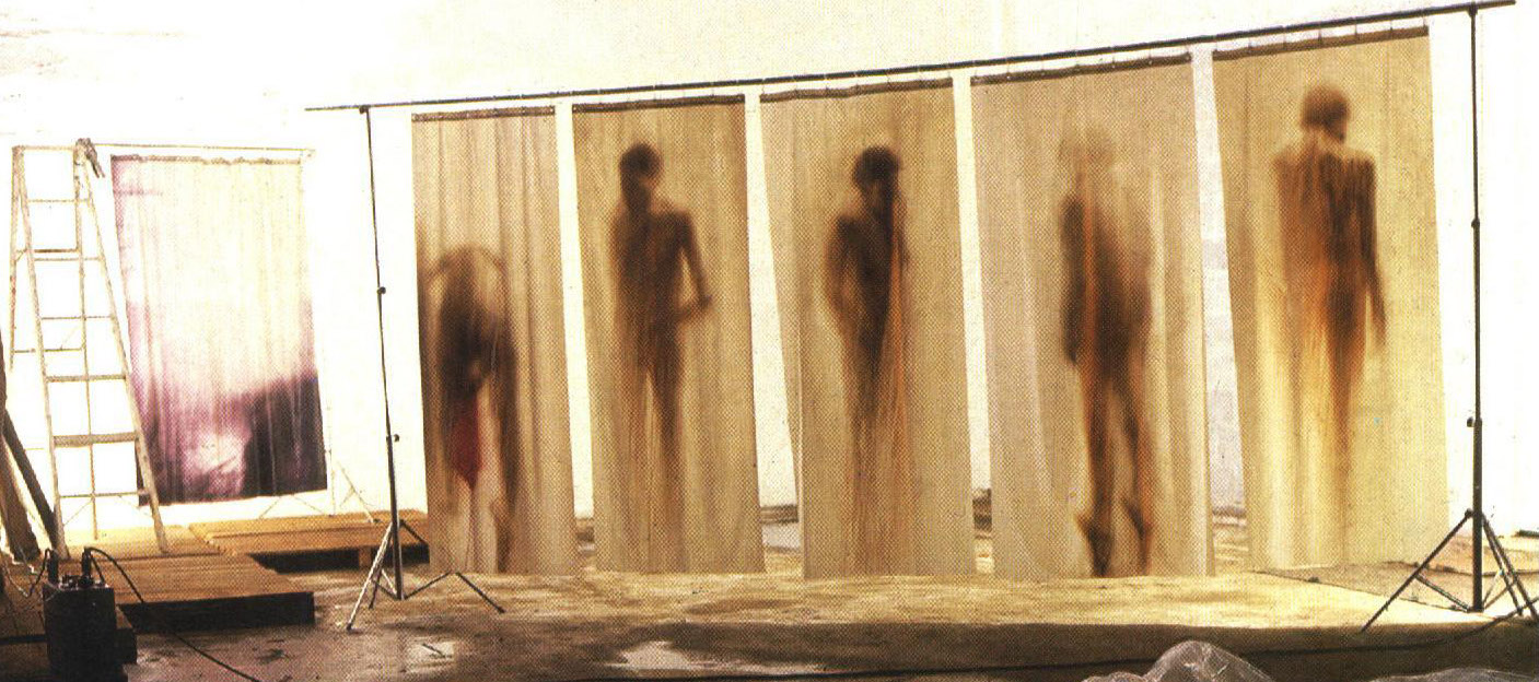 Fotografía en color de cinco cortinas de ducha, cada una de las cuales representa un cuerpo humano de pie, en tonos sepia. A la izquierda, una escalera. En primer plano, el suelo tiene un poco de agua que refleja las imágenes de los cuerpos humanos.