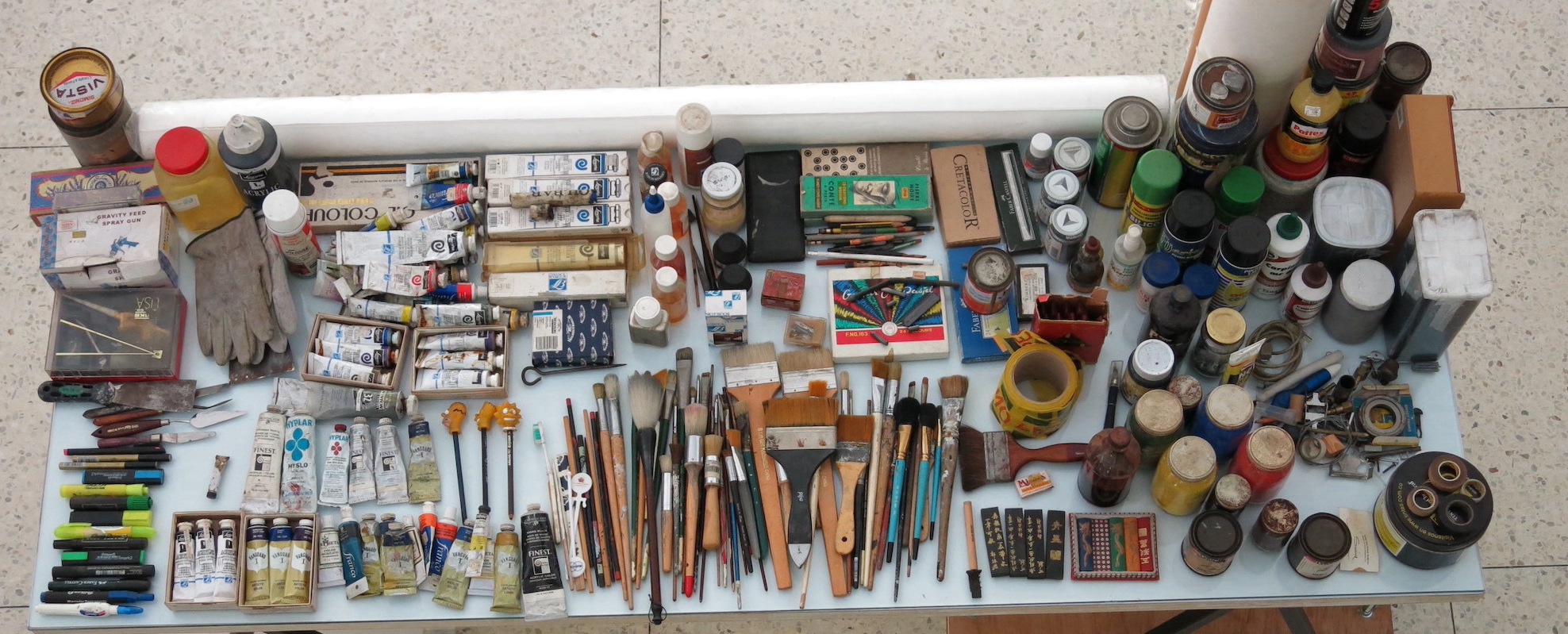 Una mesa vista desde arriba. En la superficie de la mesa, hay lápices, pinceles, pinturas y diferentes materiales y herramientas usados por artistas.