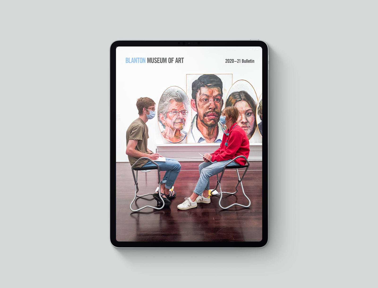 An iPad with an image of two people facing each other while sitting on stools in front of large portraits inside a gallery.