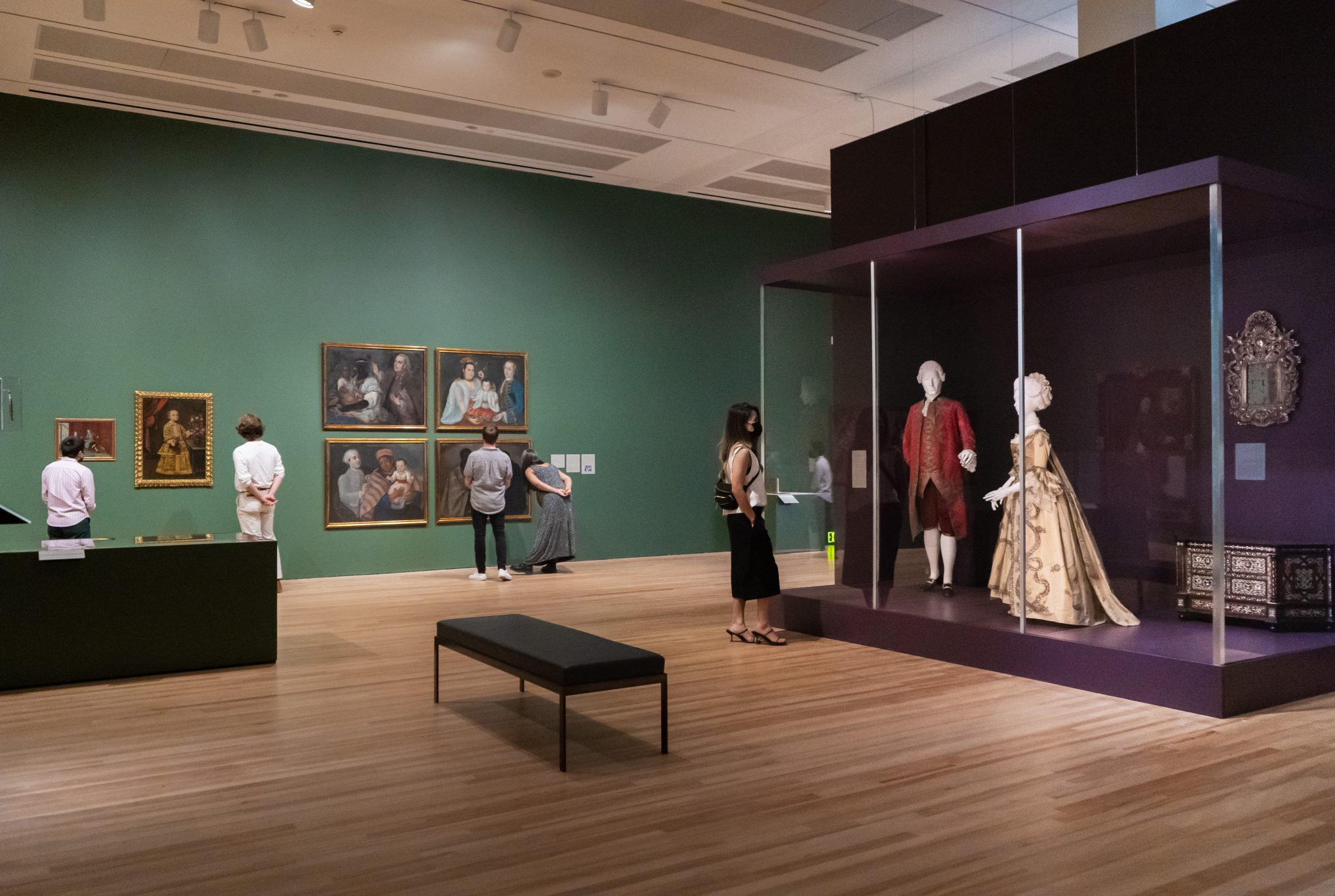 An installation view of an exhibition featuring paintings on a wall and two mannequins wearing 18th-century formalwear inside a glass enclosure.