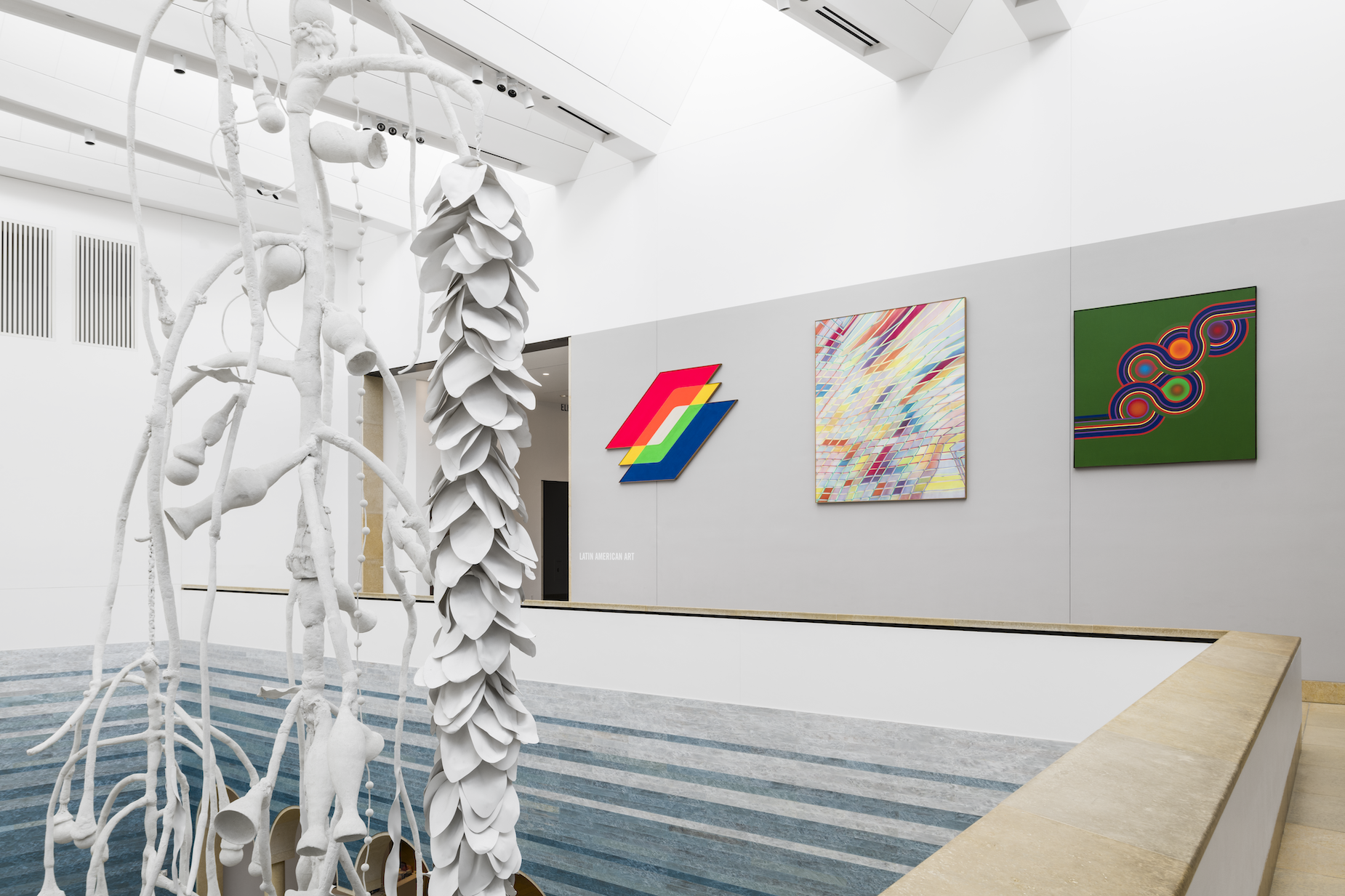The mezzanine of a museum with 3 large abstract works on the wall in the background. In the foreground is a large, dangling white sculpture with oceanic qualities. It hangs over the atrium which is covered with gradient blue-colored tiles
