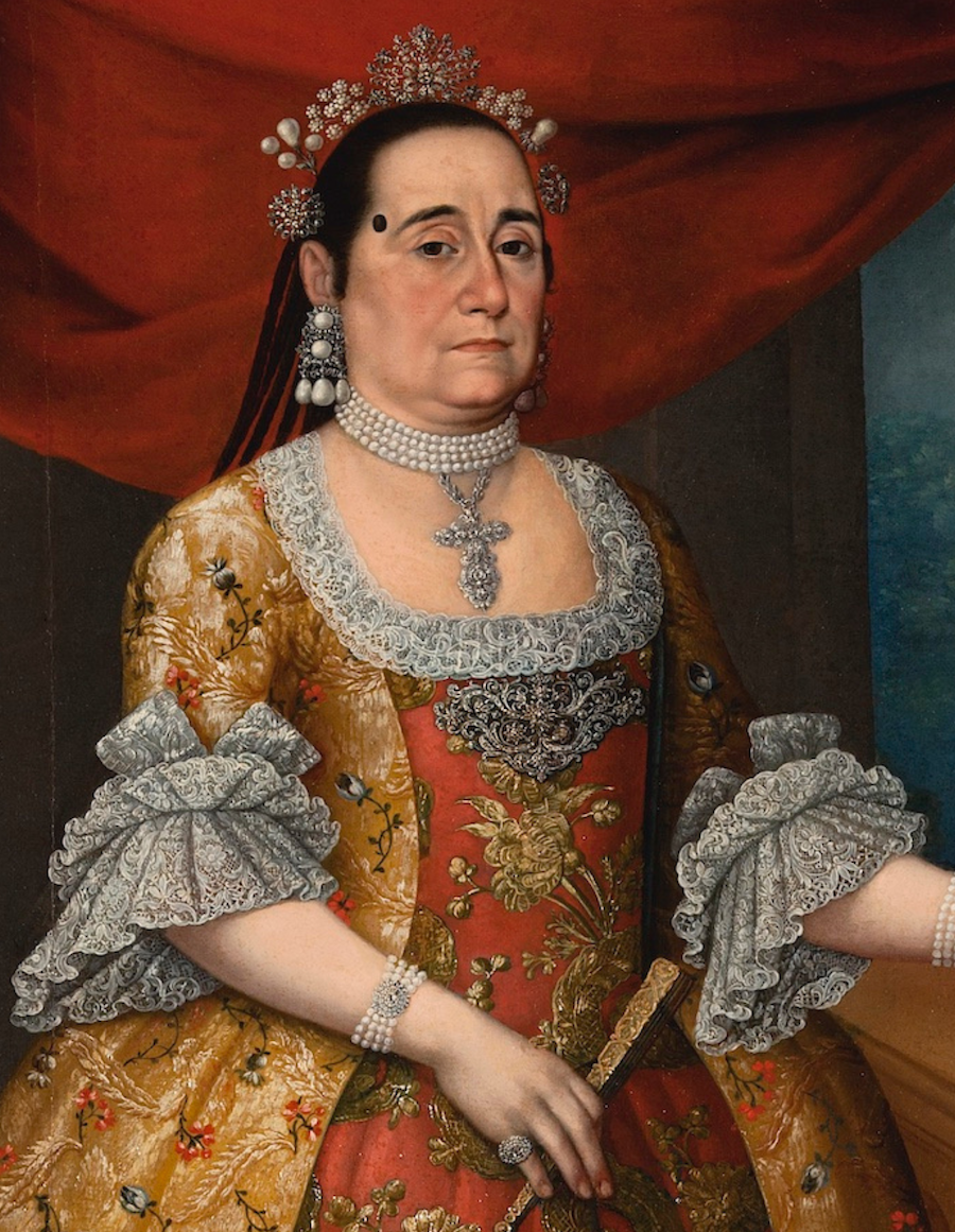 A woman wearing an ornate 18th-century dress, standing in front of a red curtain.