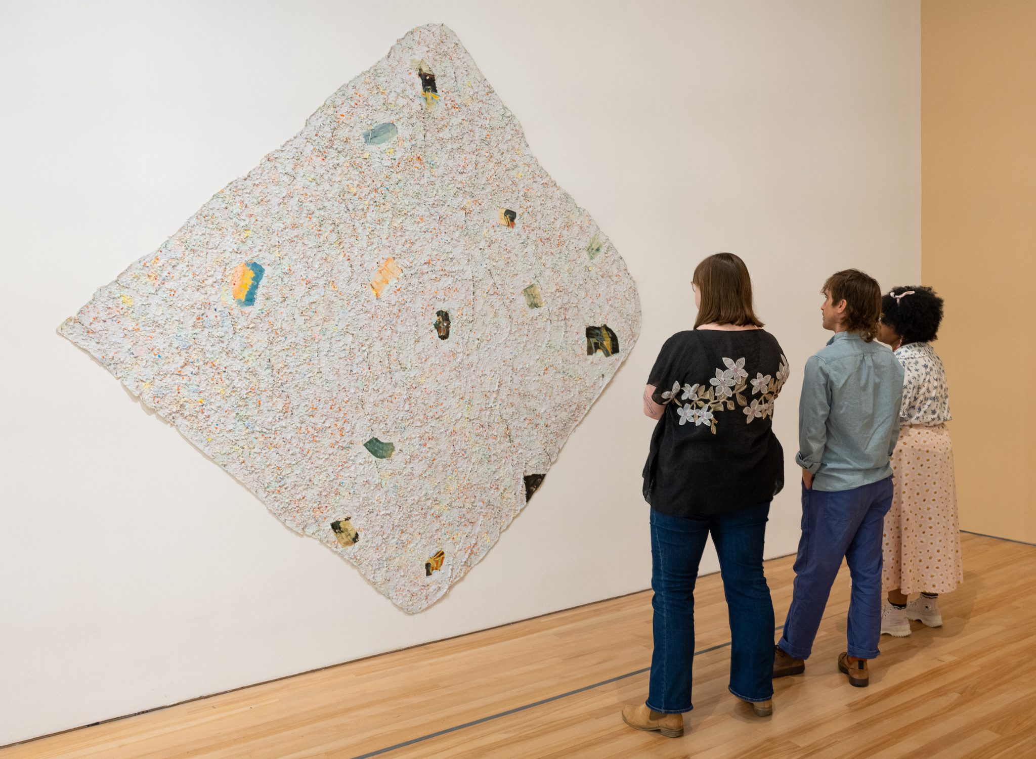 3 people standing in front of a large abstract artwork with lots of punched paper holes over the canvas