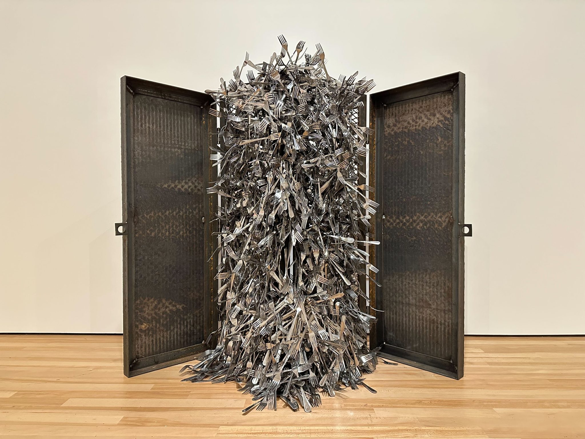 A metal artwork constructed of 3 panels. The center panel is completely covered with metal forks which are stuck on magnets
