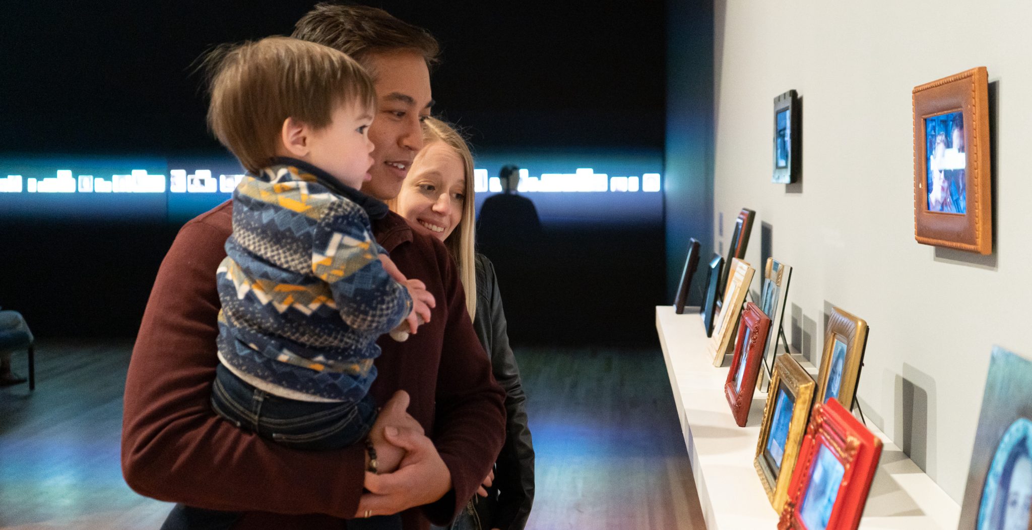 A family of three looking at some picture frames on a shelf inside an exhibition space