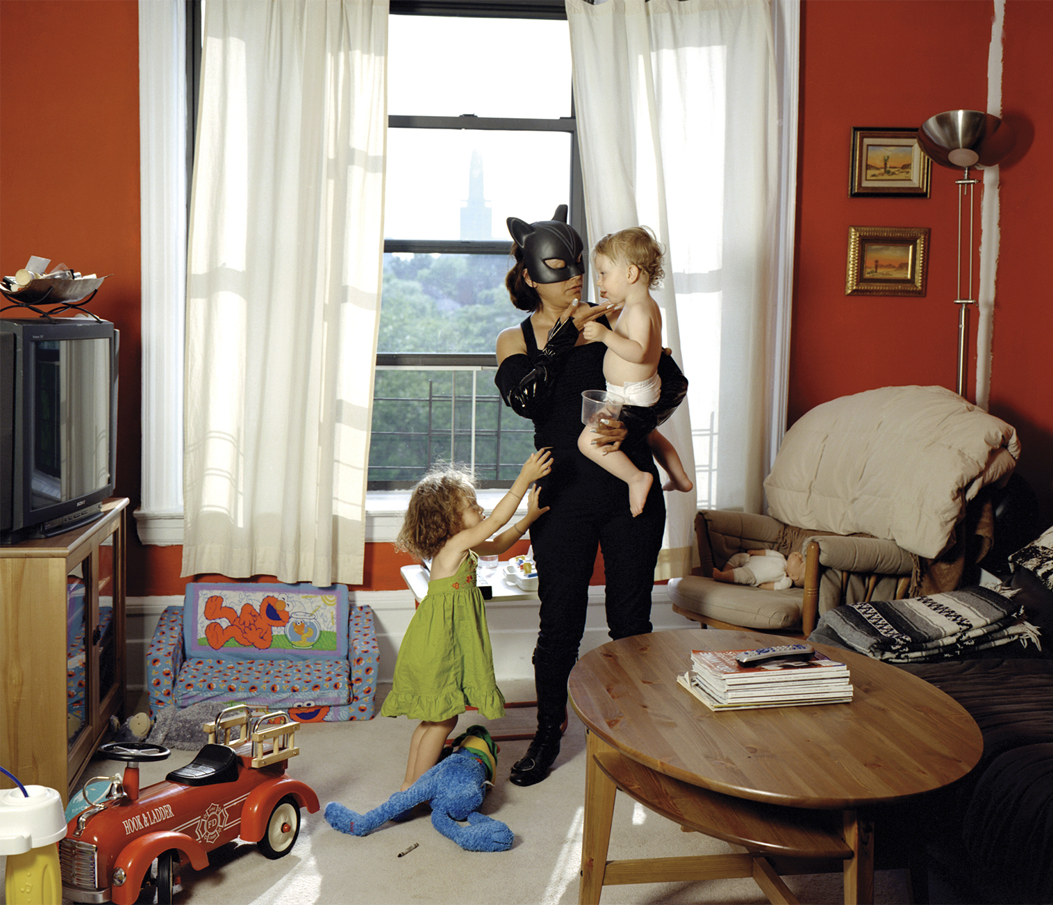 A woman dressed as Catwoman holding a baby inside an apartment. A toddler wearing a green dress pulls at her