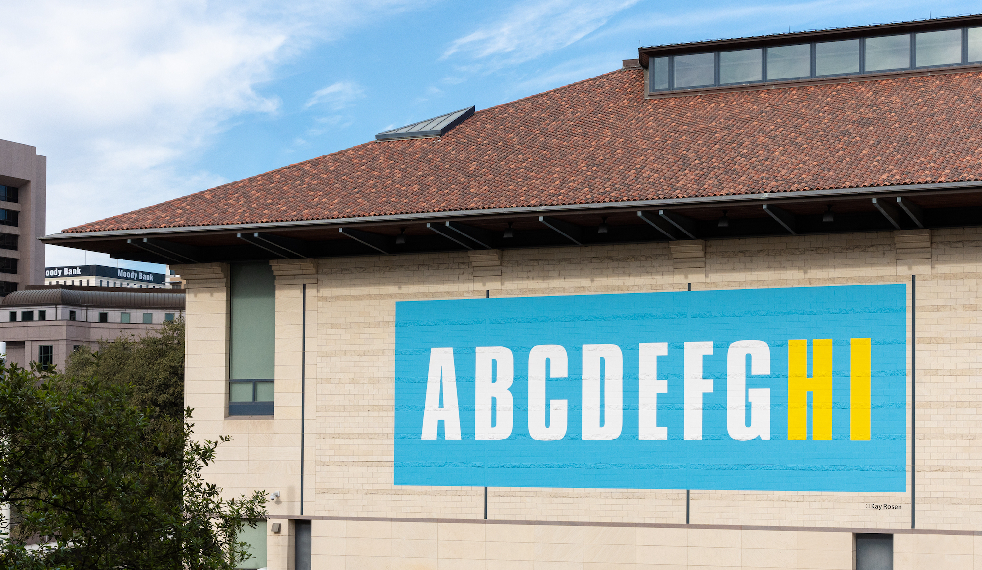 A large blue mural at the side of a large museum building with ABCDEFG in white letters and HI in yellow letters