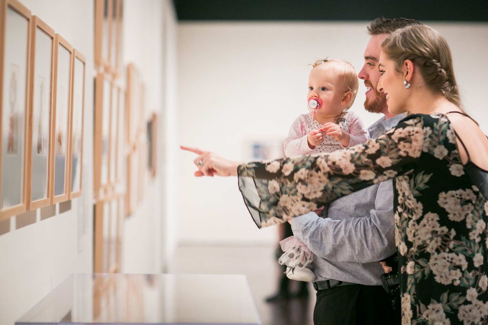 A couple holding a baby, looking at and pointing toward works of art in a gallery