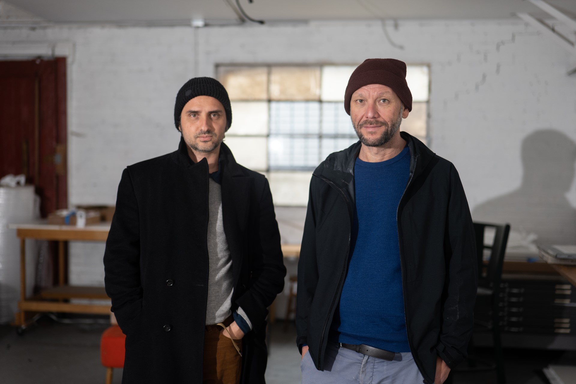 Two men wearing black jackets and beanie hats, standing inside an artist studio