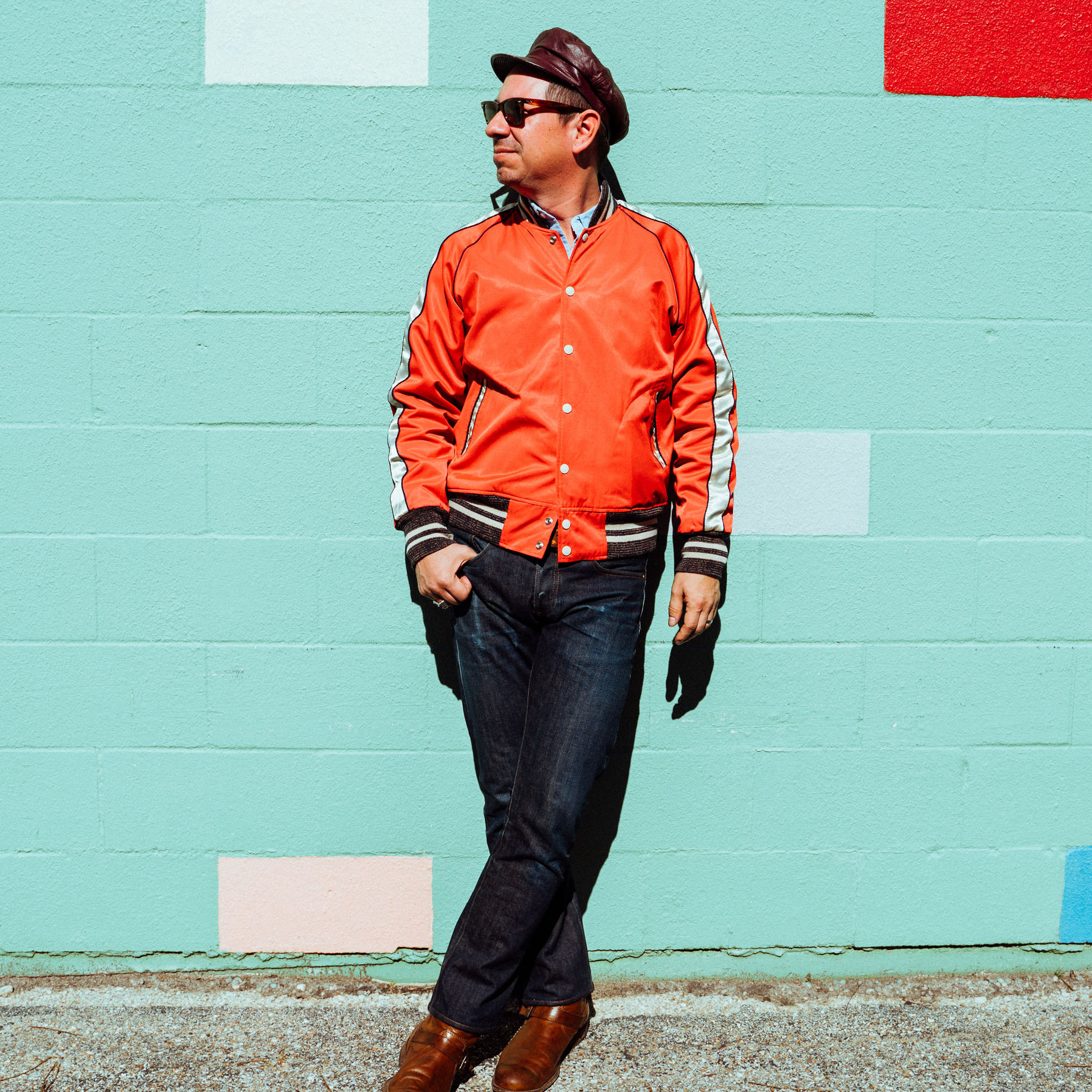 A man wearing dark trousers, an orange jacket, sunglasses and a hat, leaning against a light blue wall