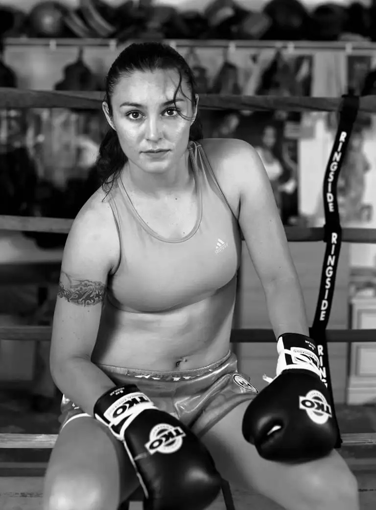 An unsmiling young woman sitting with her hair tied back while wearing boxing gloves, shorts, and a sports bra.