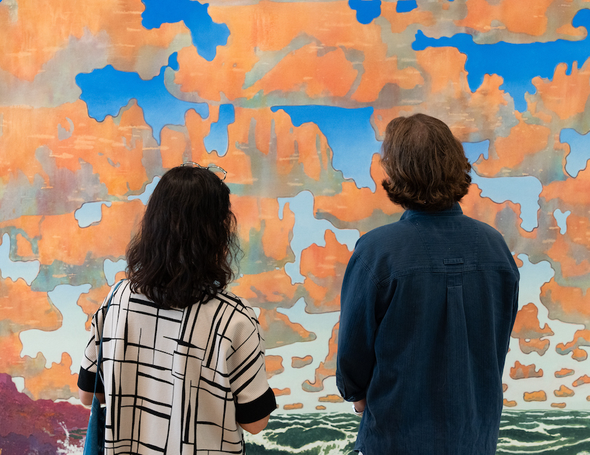 Two people looking at a large painting with orange clouds over a blue sky