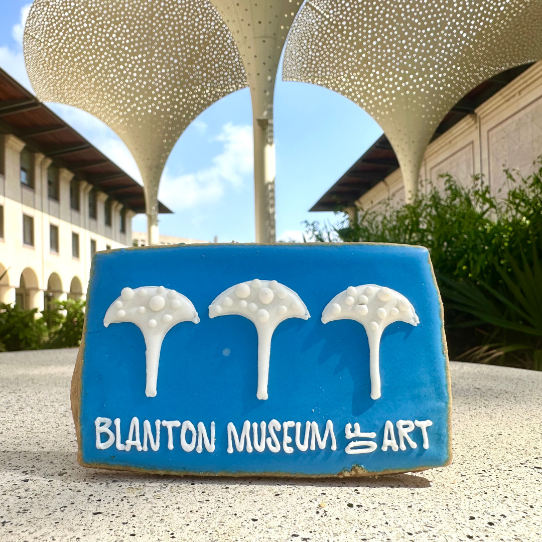 A blue cookie with 3 petal-like structures and text reading "Blanton Museum of Art" on the front, situated on a stone surface with the grounds of the Blanton Museum in the background