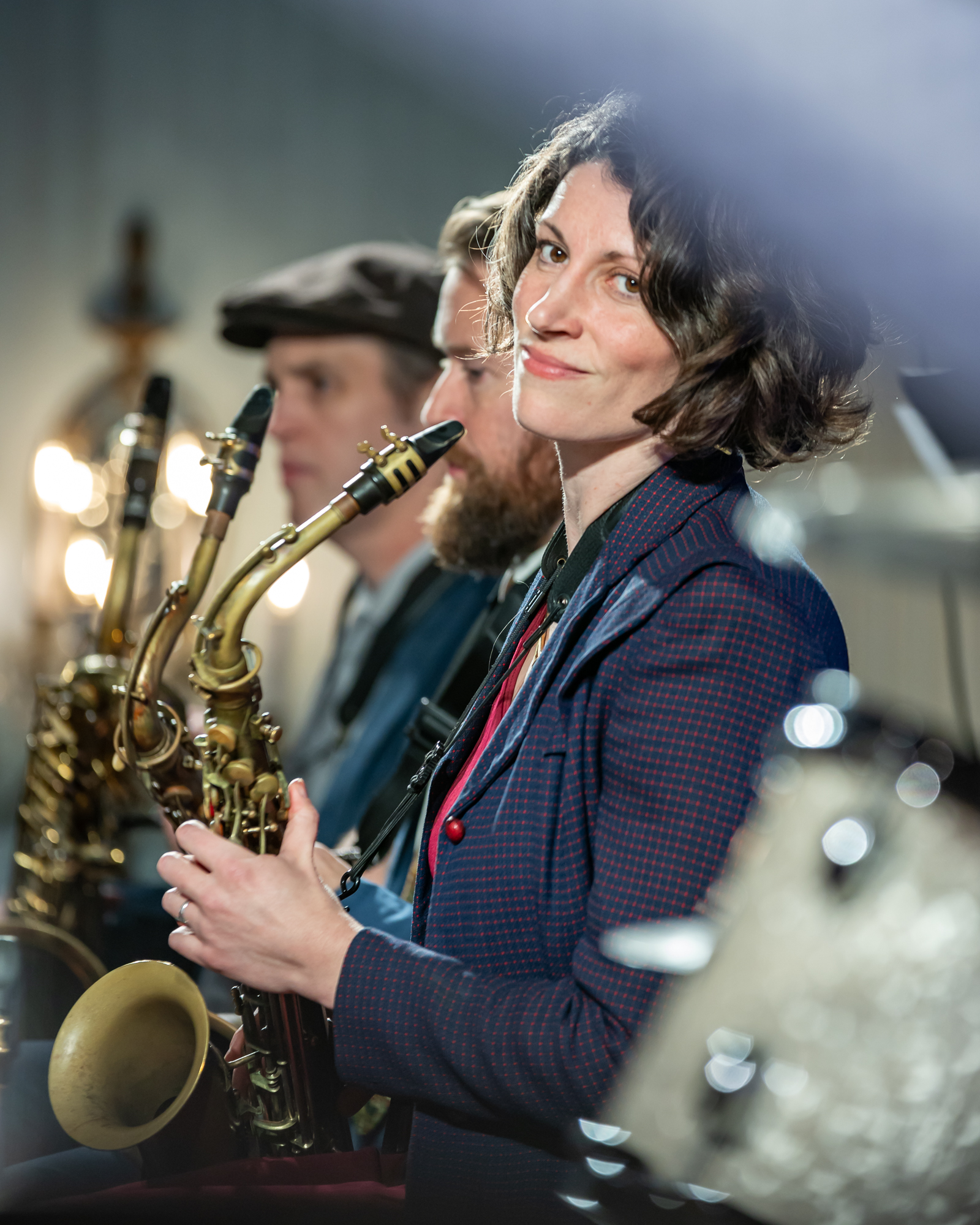 A woman holding a sax looking toward the camera with other brass instrument players sat behind her