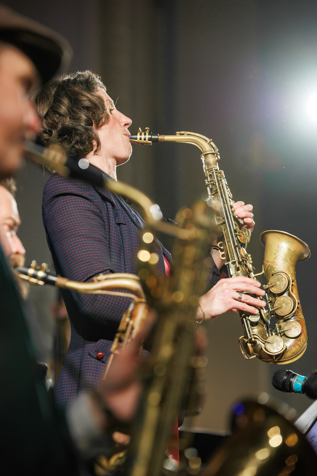 A jazz band performs on stage, featuring a saxophonist in focus playing intensely with others in the background partially visible, instruments in hand. Bright stage lighting enhances the vibrant scene of the musical performance.