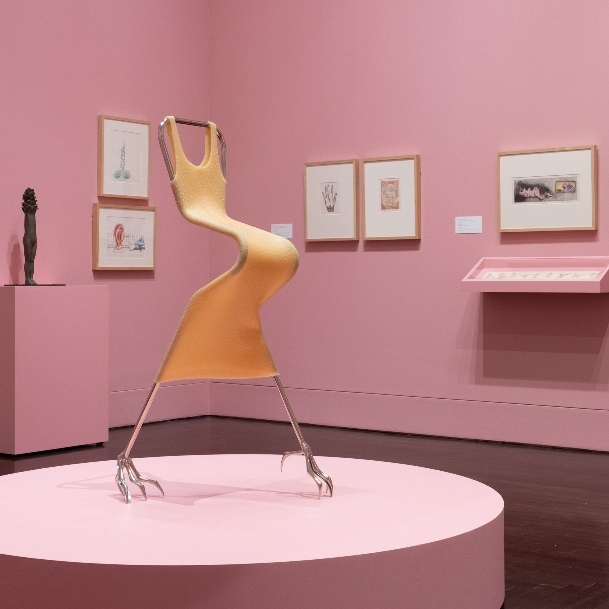 A gallery installation of Surrealist artworks with a chair-like structure on a round platform with chicken feet as legs and covered in silicone. The surrounding walls are also pink