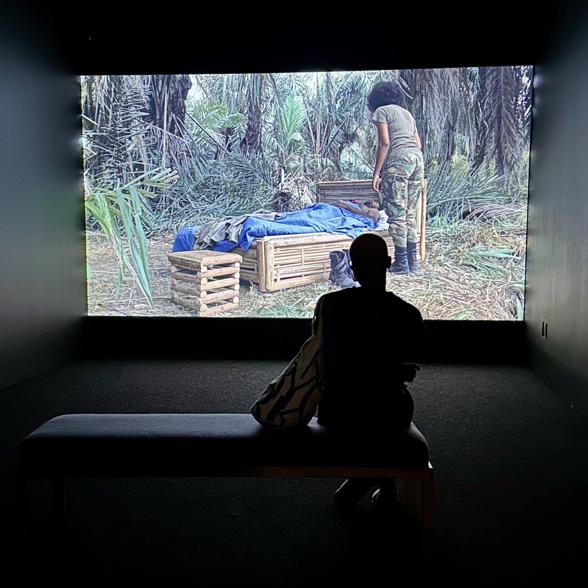 A person on a bench watching an art film featuring a woman in a rainforest setting, standing next to a bed