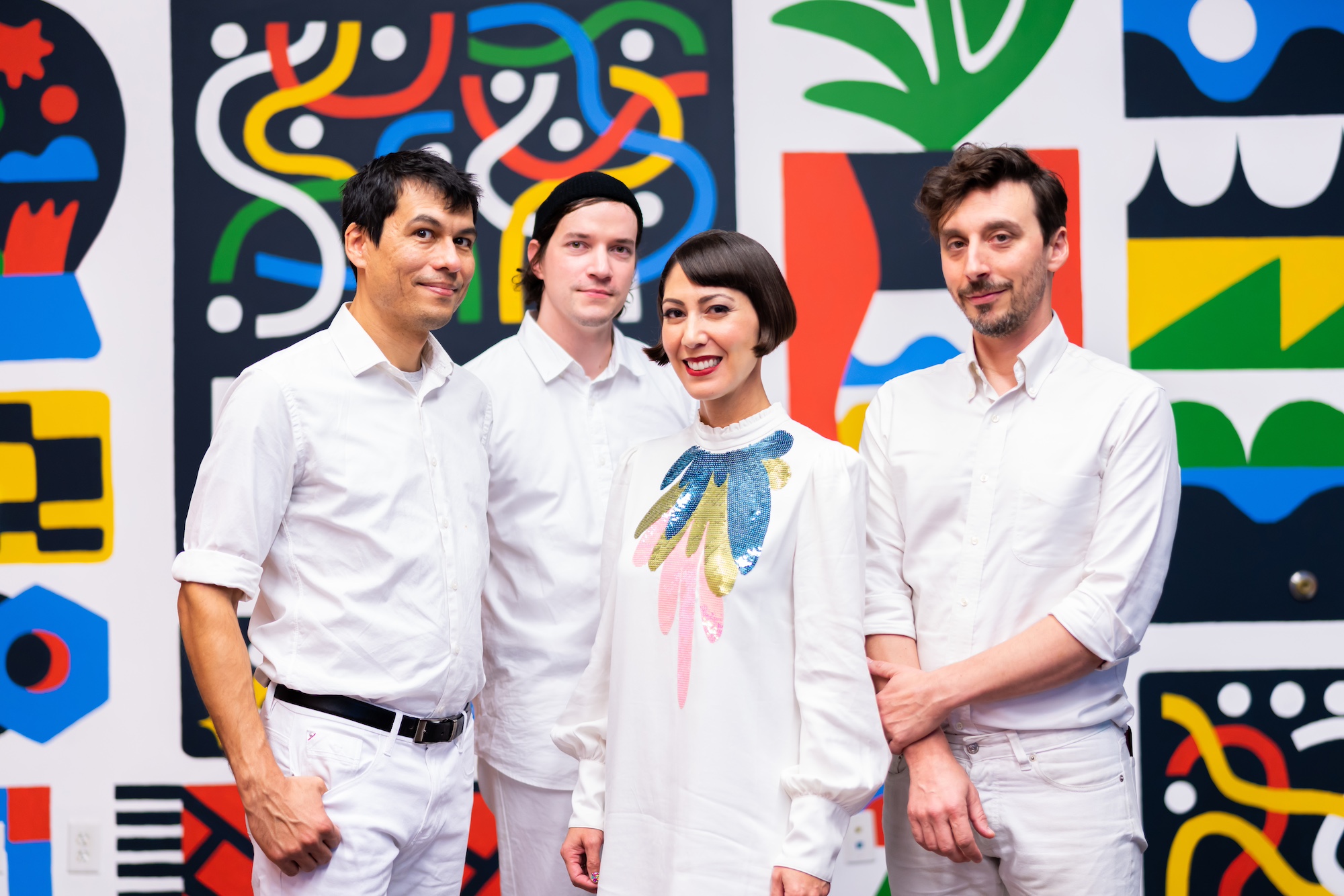 4 people from the band Octopus Project. They are all wearing white outfits and smiling, while standing in front of a vivid, graphic background of different patterns