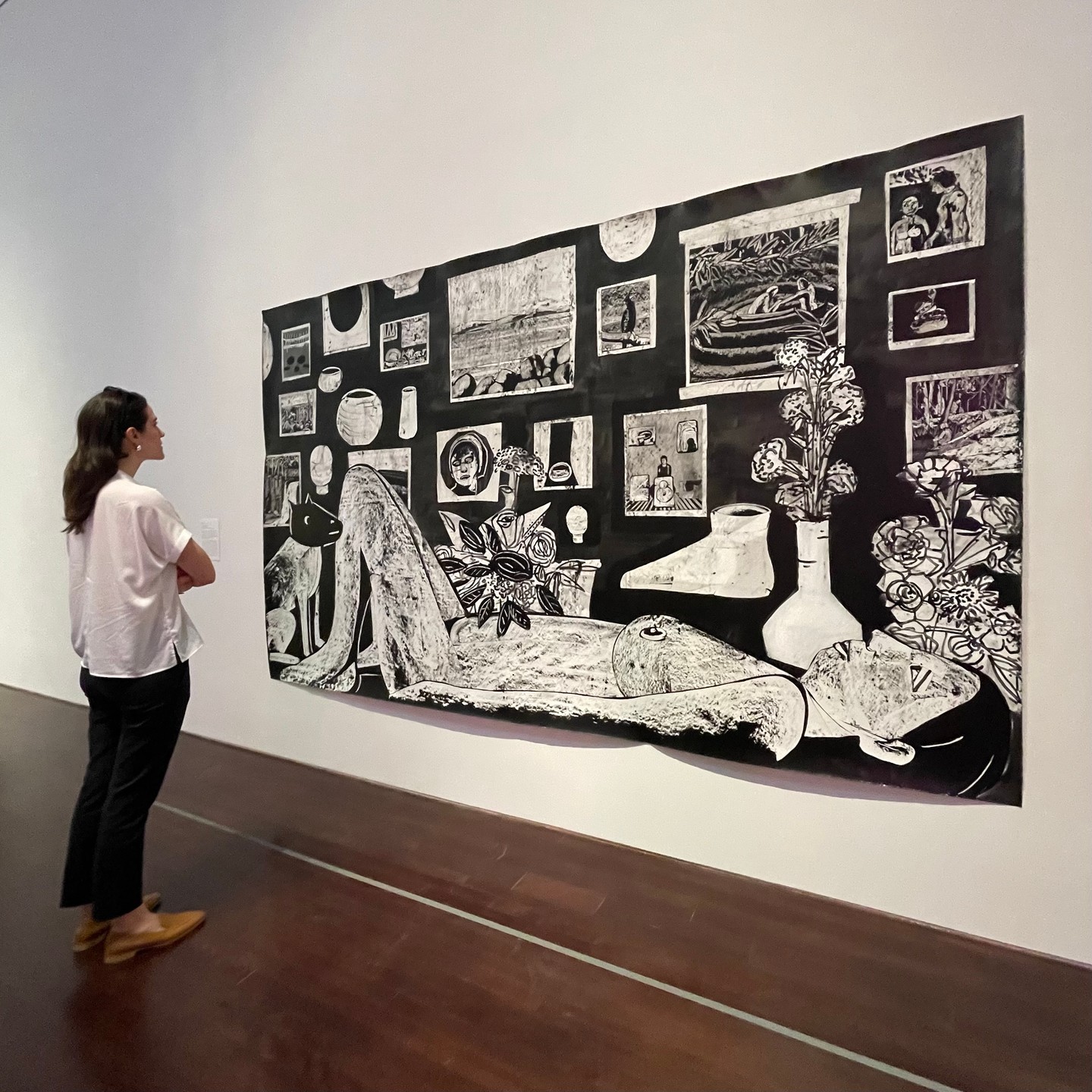 A woman looking at a large black and white artwork depicting a woman lying down in a doctor's office, surrounded by artworks on the wall and plants